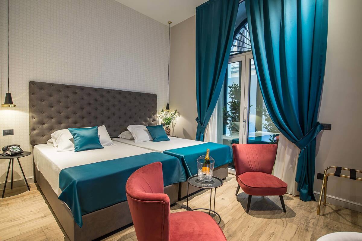 A hotel room with a large bed, two red chairs, a small table with a bottle, and a view of the outdoors through a window with blue curtains.