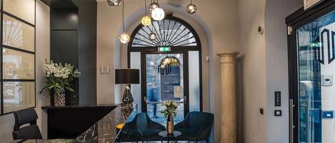 A lobby with a reception desk, two blue armchairs, a vase with flowers, a geometric patterned floor, and a large arched window.