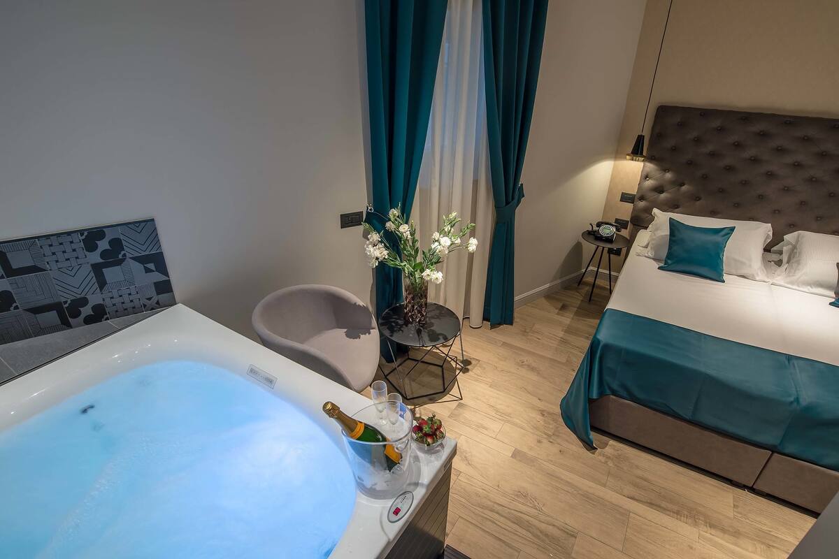 A modern hotel room with a bathtub, a bed with teal bedding, a small table with a vase of flowers, and a nightstand with a lamp.