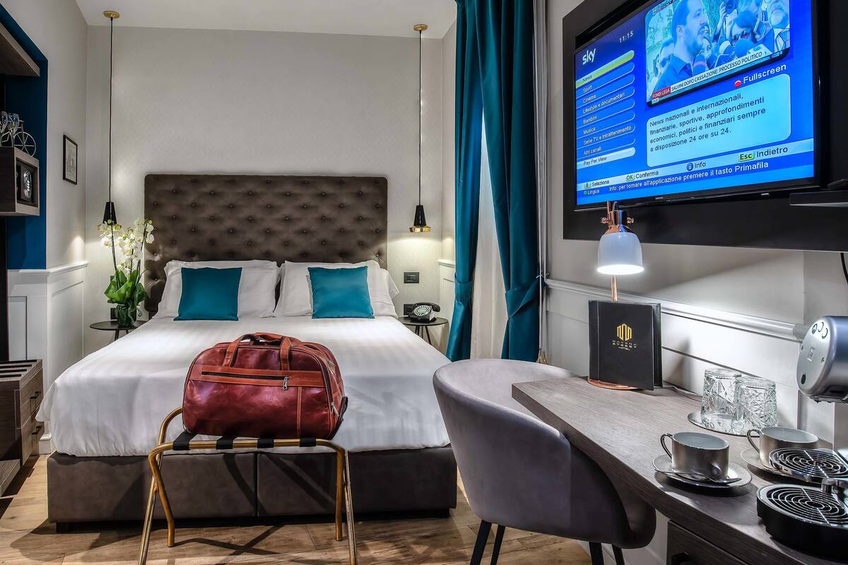 A hotel room with a bed, a desk with a coffee maker, a TV displaying a news channel, and a suitcase on a chair.