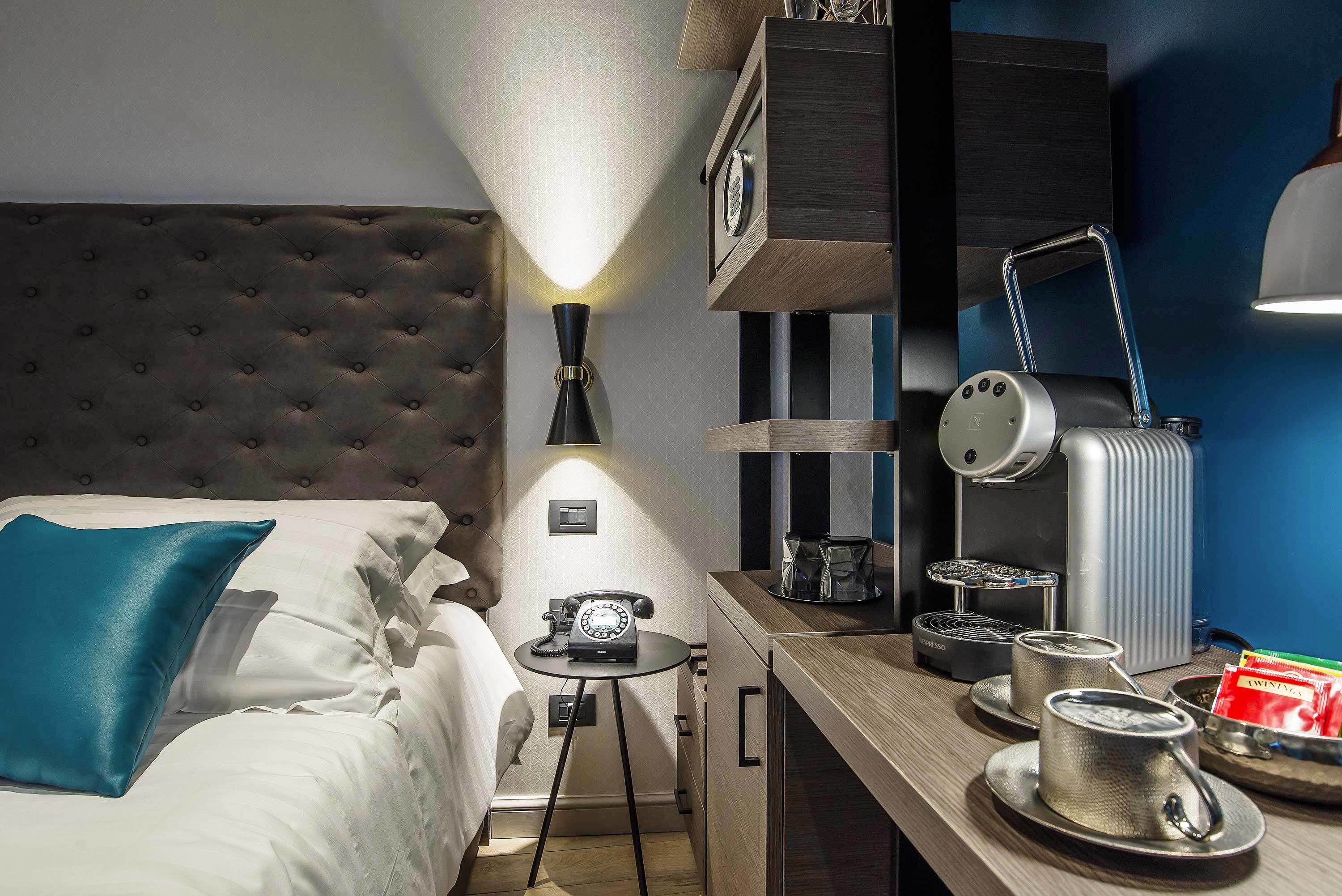 Ein Hotelzimmer mit einer gepolsterten Kopfteil, einem Bett mit weißen Bettwäsche und einem türkisfarbenen Kopfkissen, einem kleinen runden Tisch mit Telefon und Lampe, einer Kaffeemaschine und einem Tablett mit Teekannen.