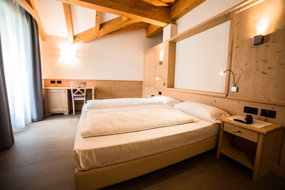 A hotel room with a wooden ceiling, a bed with white bedding, a desk with a chair, and a nightstand with a lamp.