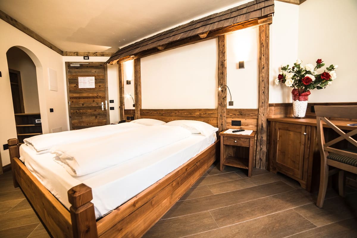 A wooden bed with a headboard, a bedside table with a lamp, a small desk, and a vase of flowers.