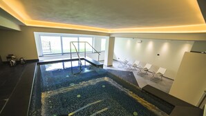 Una piscina interior moderna con suelo de cristal, una escalera metálica y un área de descanso con sillones.