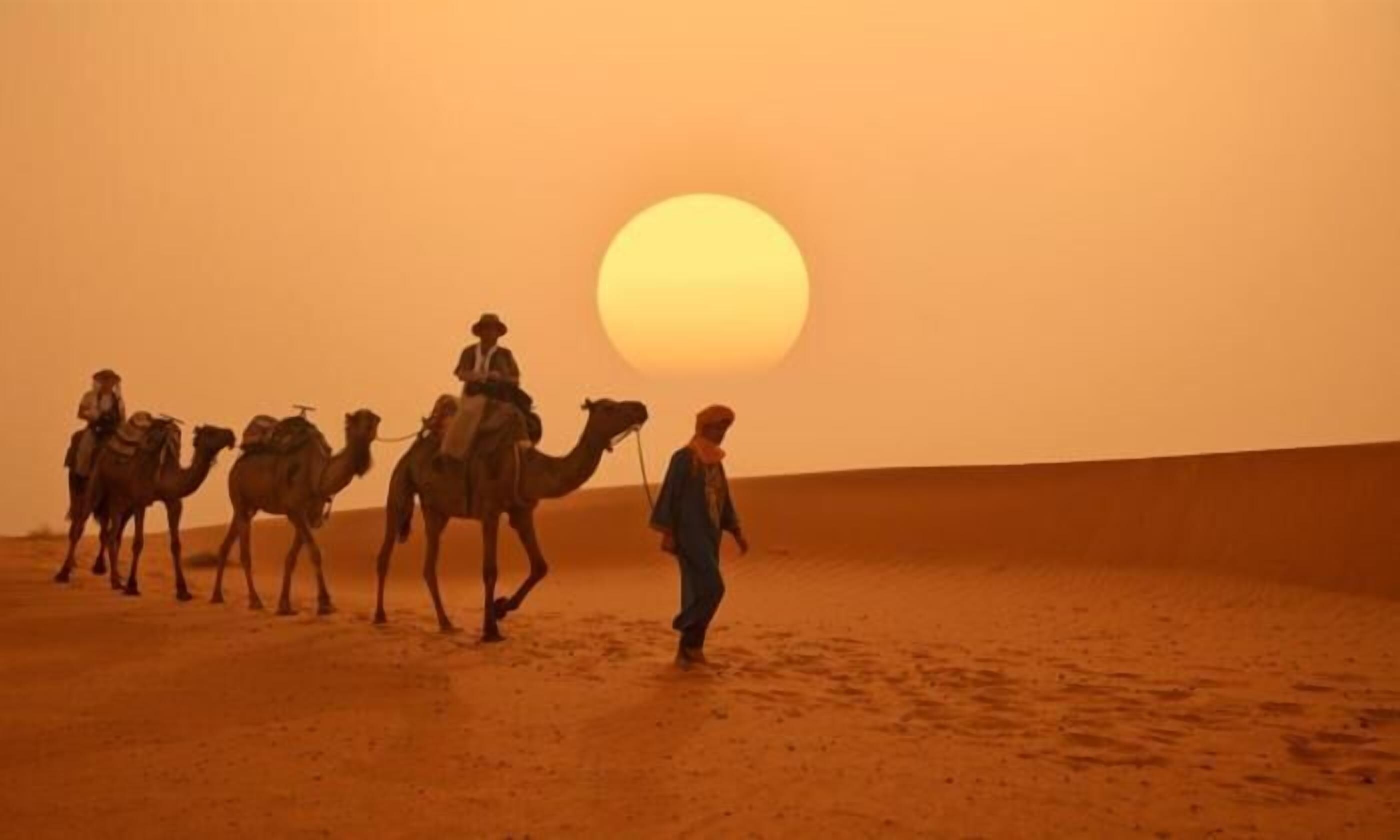 A desert landscape with a large sun in the sky and camels with riders.