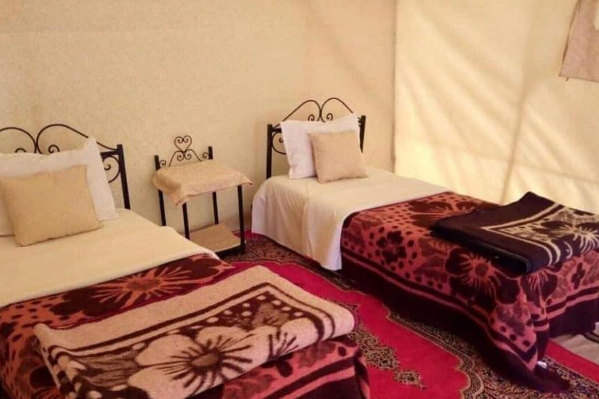 Deux lits simples avec des têtes de lit, une petite table et un tapis à motifs dans une pièce qui ressemble à une tente.
