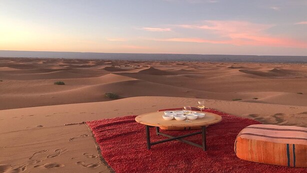 Paisaje desértico con dunas de arena y una mesa dispuesta para una comida, con cuencos y copas de vino.