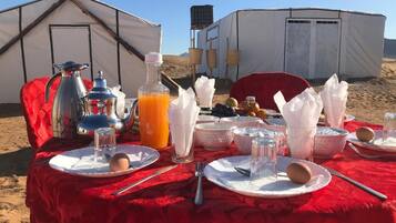A dining setup with a red tablecloth, plates, cutlery, and a beverage pitcher, set against a backdrop of tents and a water tank.