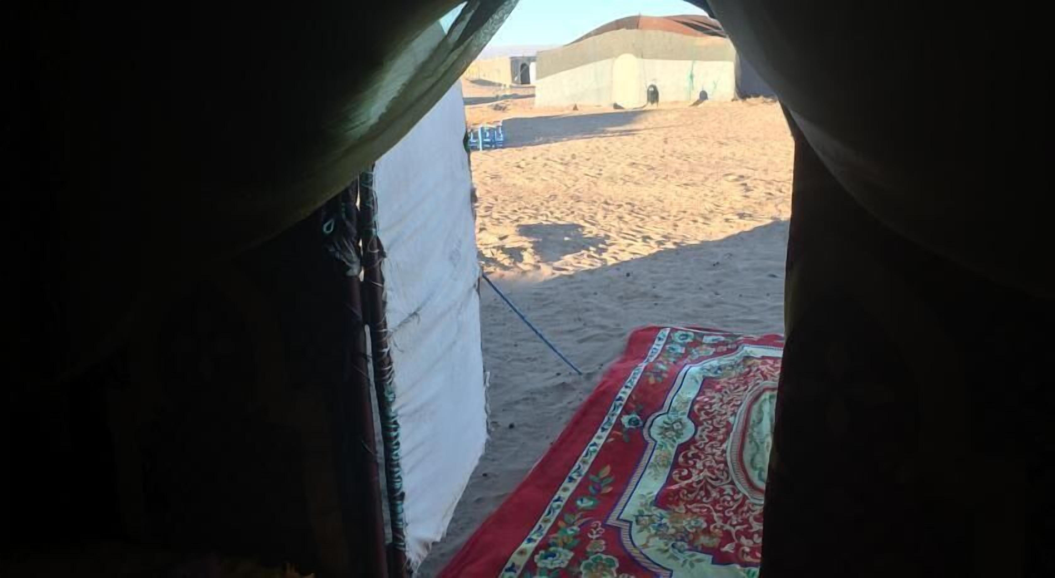 Vista desde el interior de una tienda de campaña con un tapete estampado en el suelo, una pared de la tienda y un área exterior arenosa con un edificio a lo lejos.