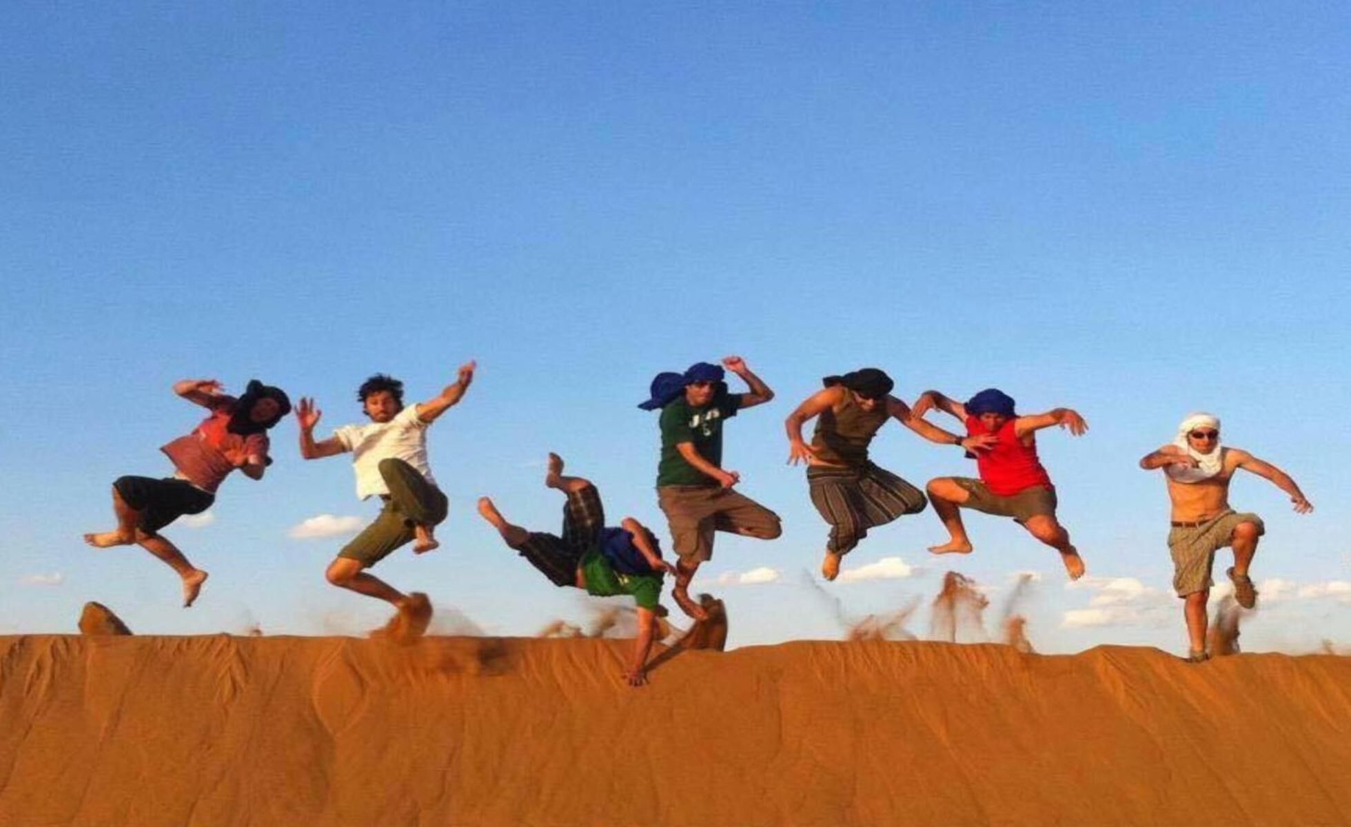 Un grupo de personas saltando en el aire sobre una superficie arenosa, con un cielo azul despejado al fondo.