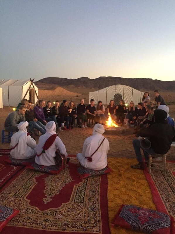 A group of people sitting on patterned rugs around a campfire in a desert setting with tents in the background.