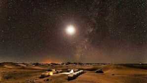 Paisaje desértico nocturno con una luna brillante y estrellas, que muestra un pequeño asentamiento con edificios y un camino de tierra.