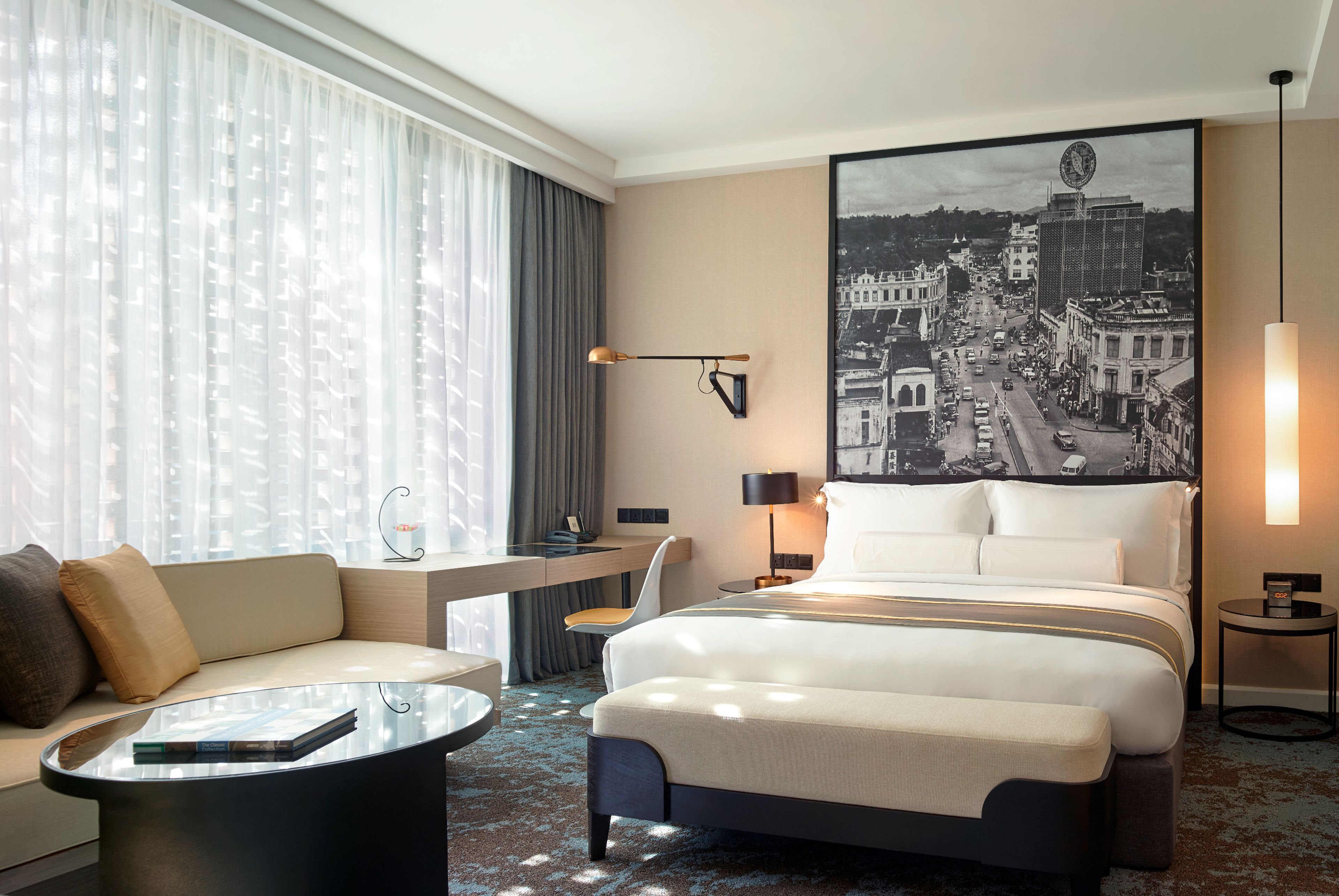A hotel room with a large bed, a desk, a sofa, a coffee table, and a cityscape painting on the wall.