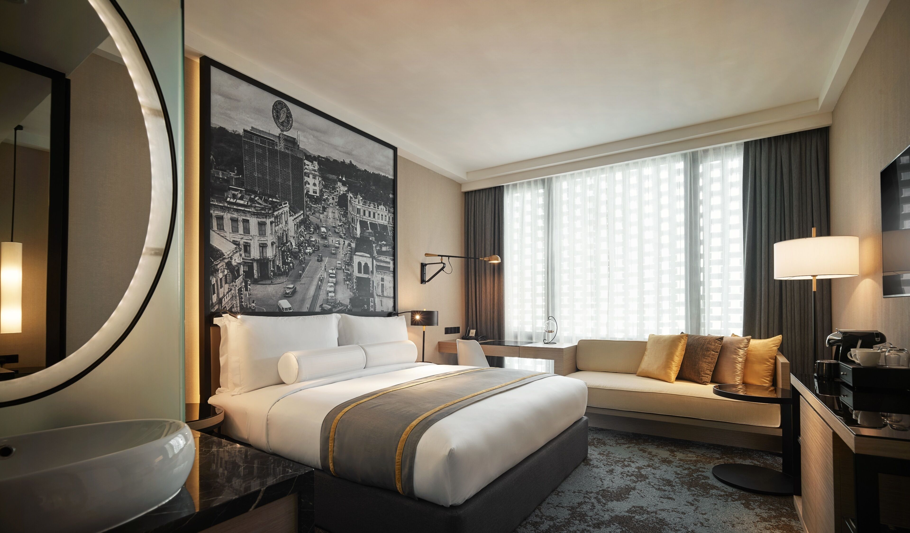 A hotel room with a large bed, a sofa, a desk, a coffee machine, and a large window with curtains.