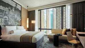 A hotel room with a large bed, a sofa, a desk, and a view of tall buildings through large windows. - Hotel Stripes Kuala Lumpur, Autograph Collection (Kuala Lumpur)