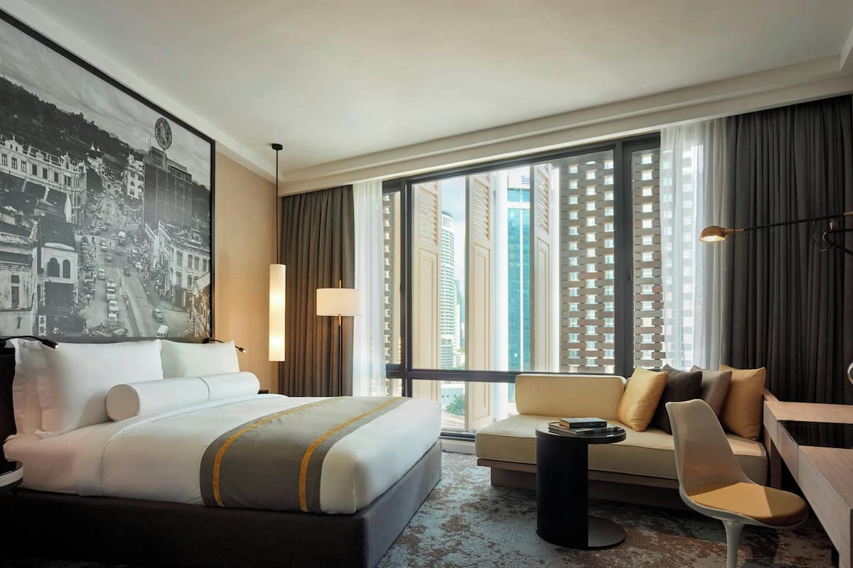 Ein Hotelzimmer mit einem großen Bett, einer Sitzecke, einem Schreibtisch und Blick auf hohe Gebäude durch große Fenster.