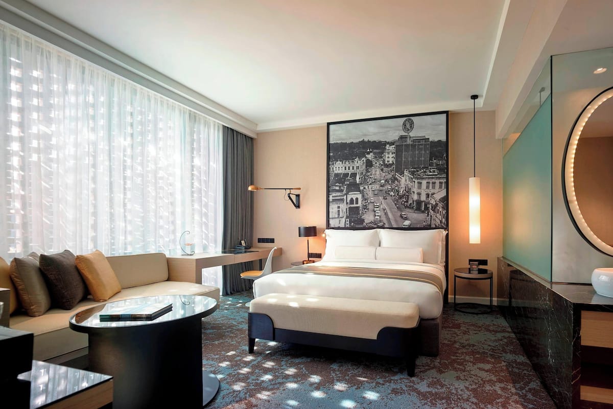 Ein Hotelzimmer mit einem großen Bett, einem Sofa, einem Couchtisch, einem Schreibtisch und einem großen Fenster mit durchsichtigen Vorhängen.