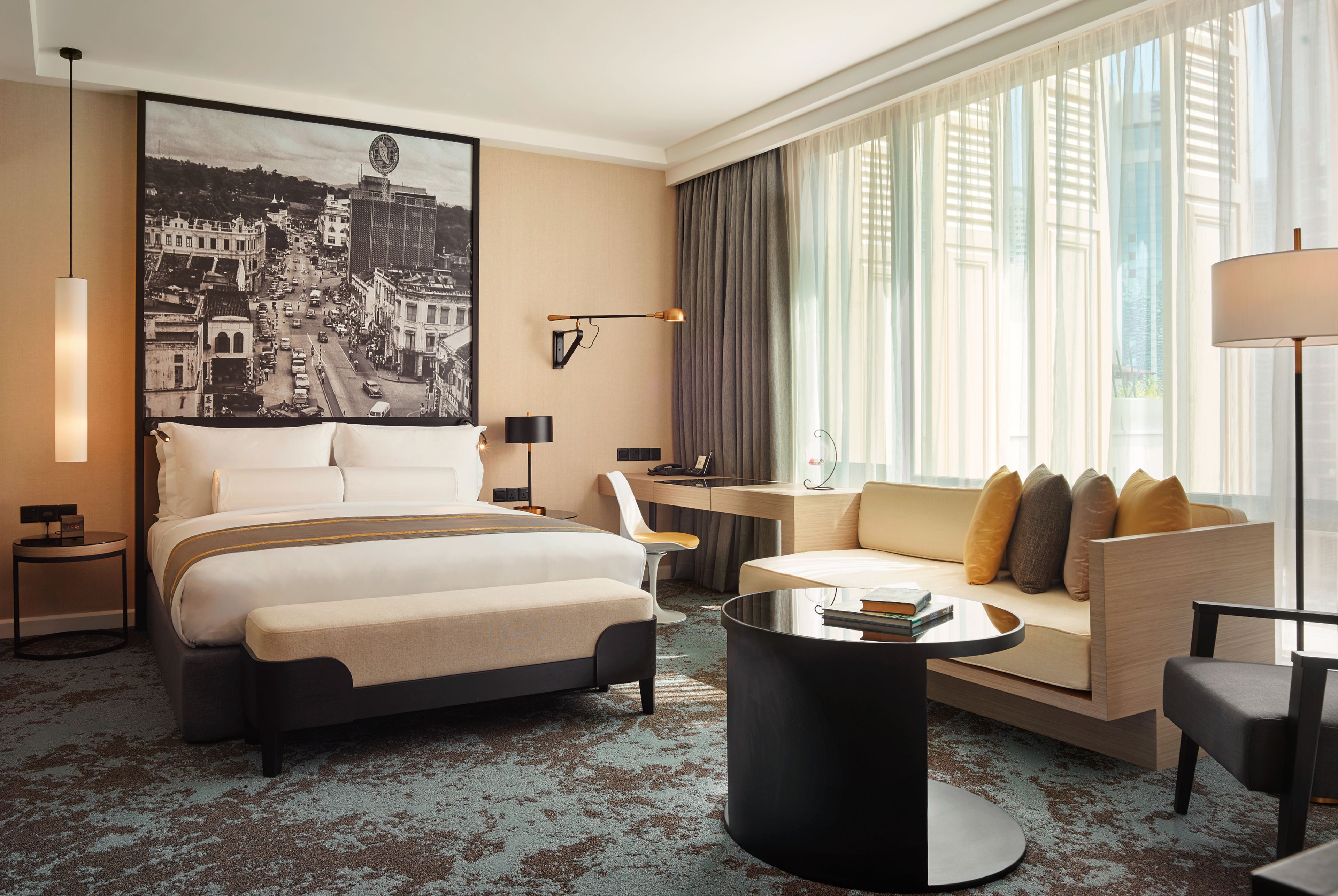 A modern hotel room with a large bed, a sofa, a coffee table, and a desk. There is a large window with curtains and a cityscape view.