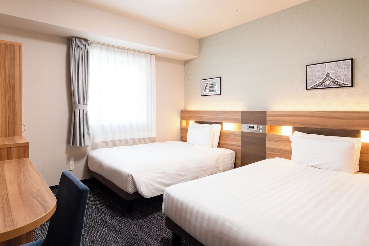 A hotel room with two beds, a desk, a chair, a TV, and a window with curtains.