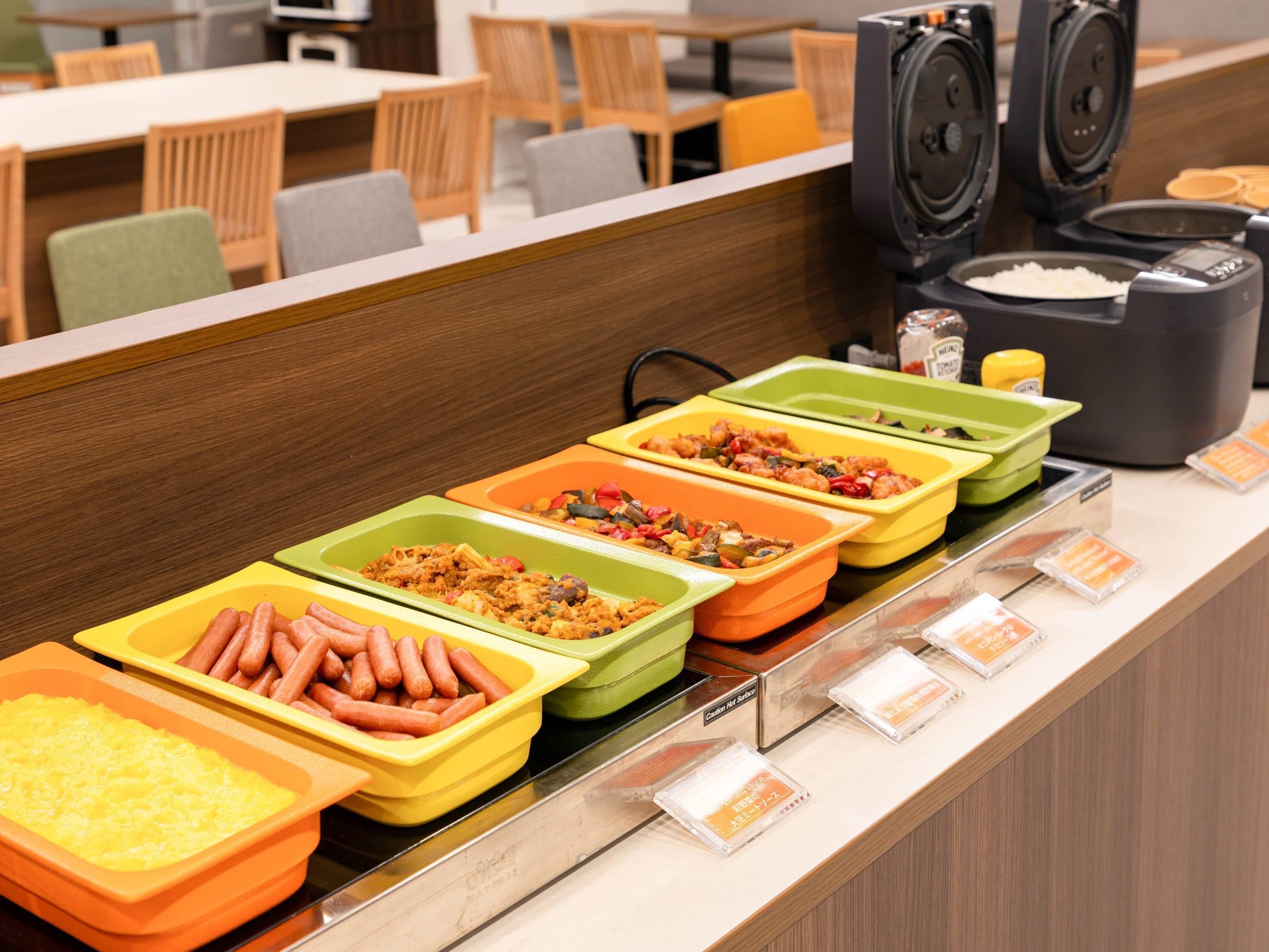 A buffet line with various food options in trays, a coffee machine, and a seating area with tables and chairs.