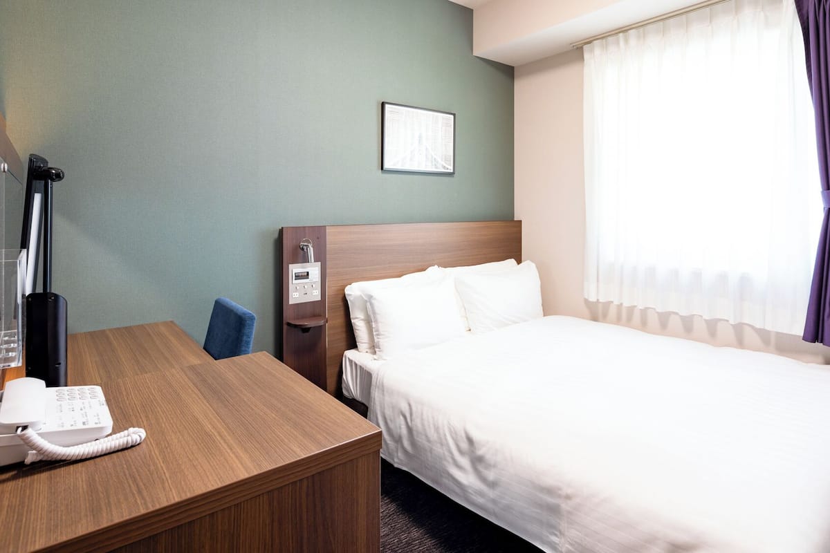 A hotel room with a bed, a desk with a telephone, a nightstand with a lamp, a window with curtains, and a framed picture on the wall.