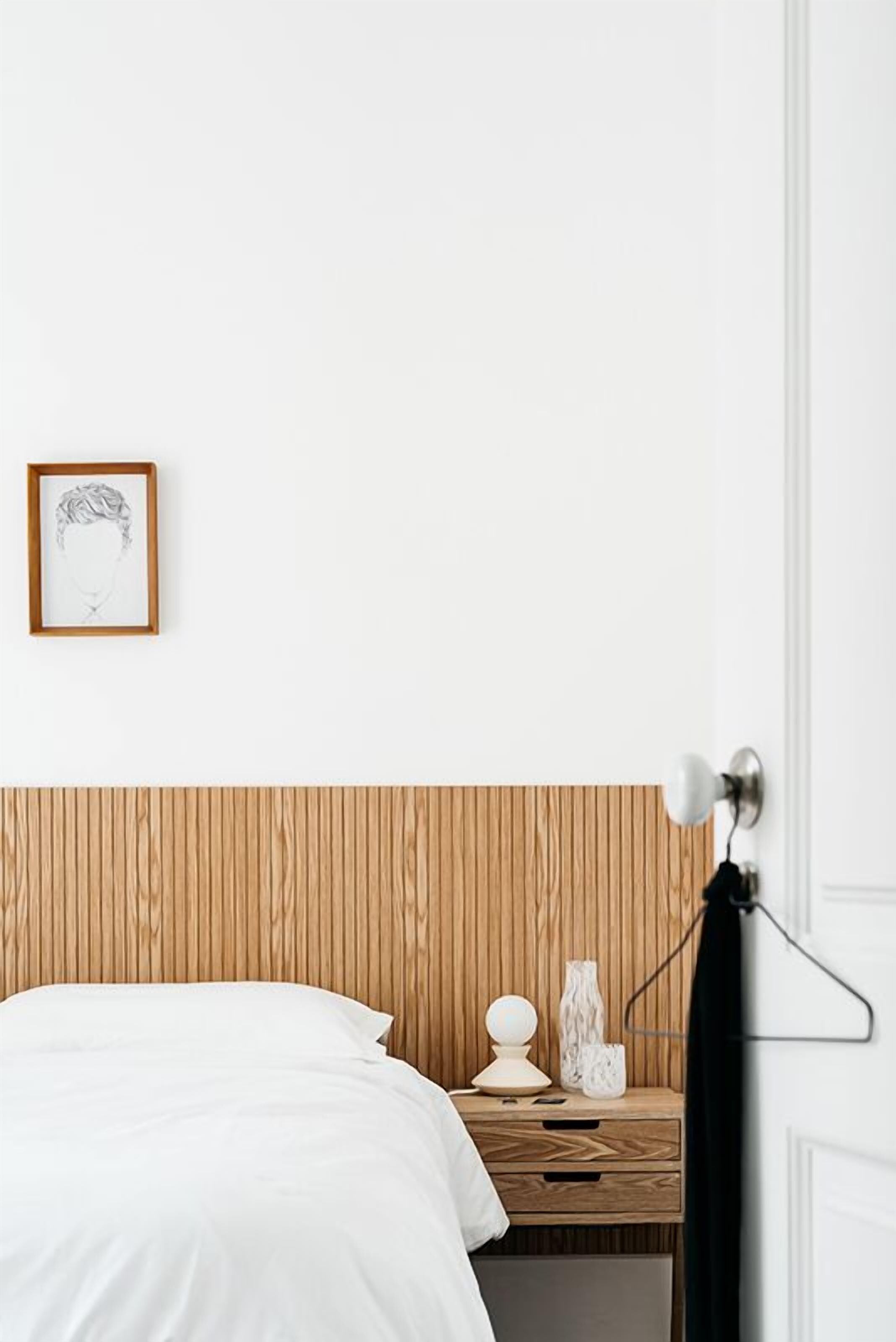 A bedroom with a wooden headboard, a bedside table with a lamp, a framed picture, and a bed with white bedding.