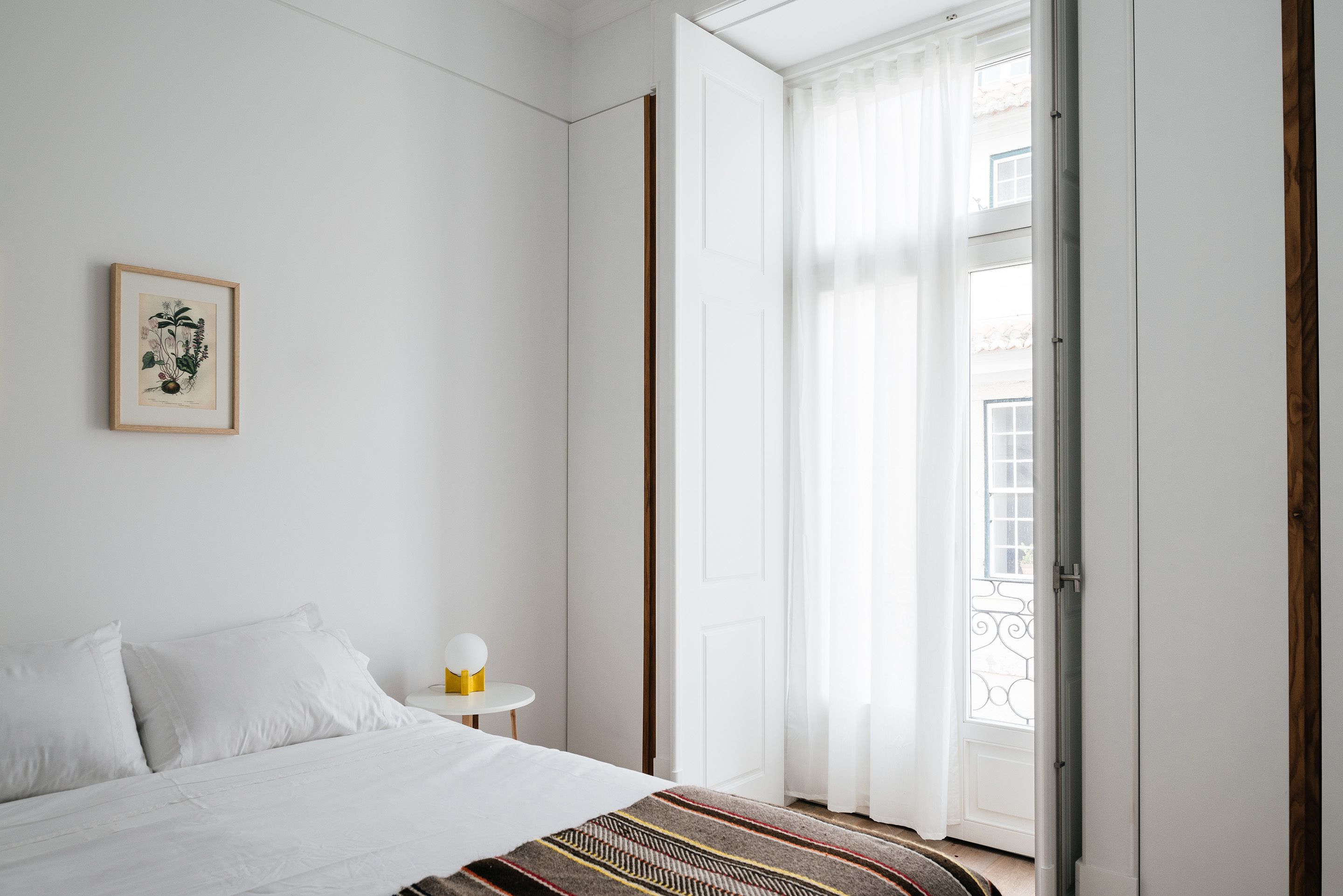 Un dormitorio con una cama, una mesita de noche con lámpara, un grabado botánico enmarco y vistas al exterior a través de una puerta.
