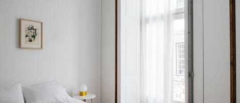 Un dormitorio con una cama, una mesita de noche con lámpara, un grabado botánico enmarco y vistas al exterior a través de una puerta.
