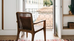 A room with a view of a courtyard, a wooden chair, a striped rug, and a bookshelf with books and a cactus.