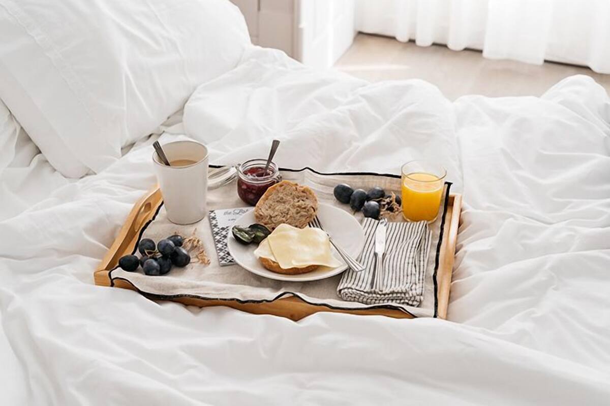 Un plateau de petit-déjeuner sur un lit, avec une tasse de café, un verre de jus d’orange, une assiette contenant un croissant, du fromage et un pot de confiture.