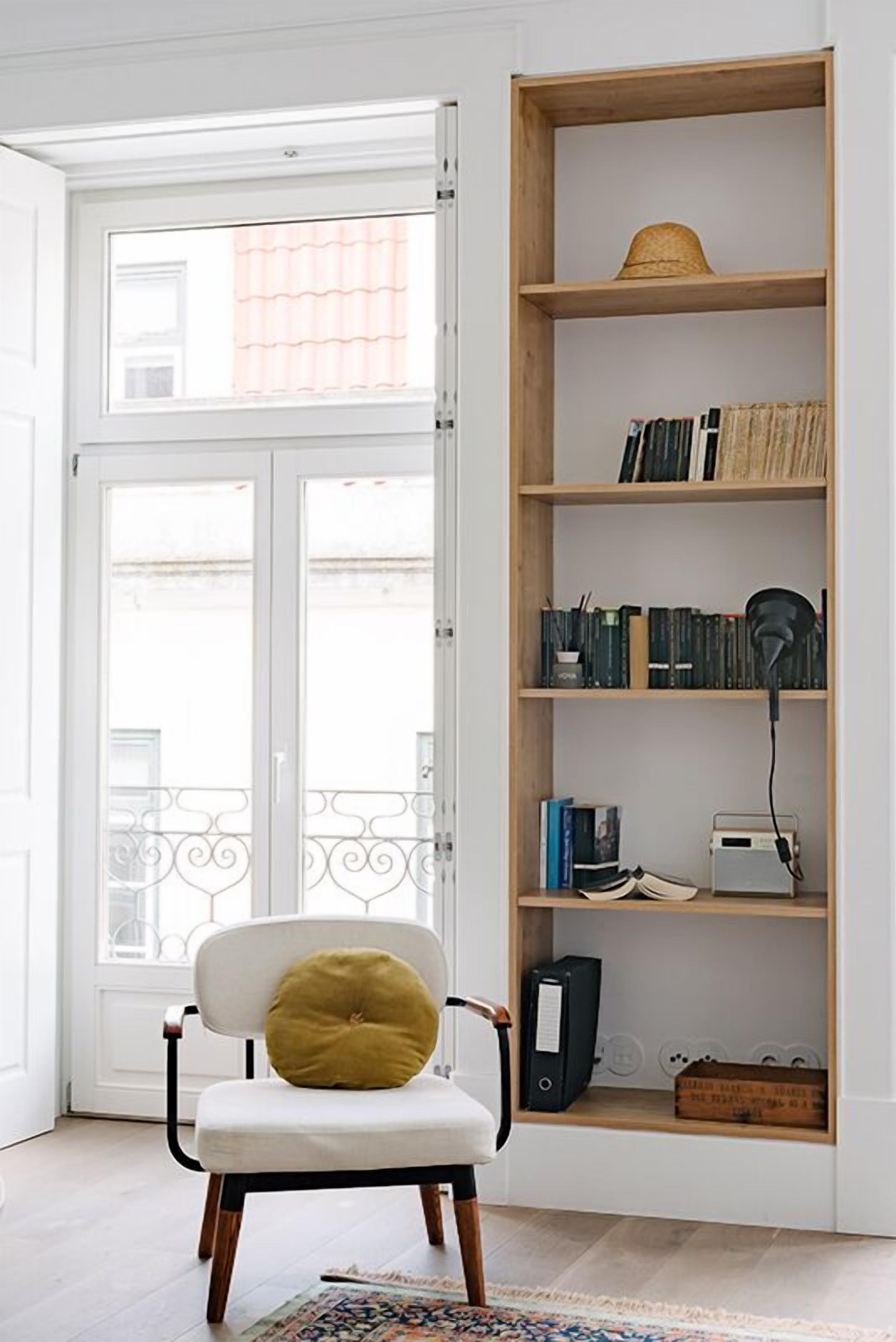 A room with a white chair with a yellow cushion, a wooden bookshelf with books and a hat, and a white door with a balcony view.