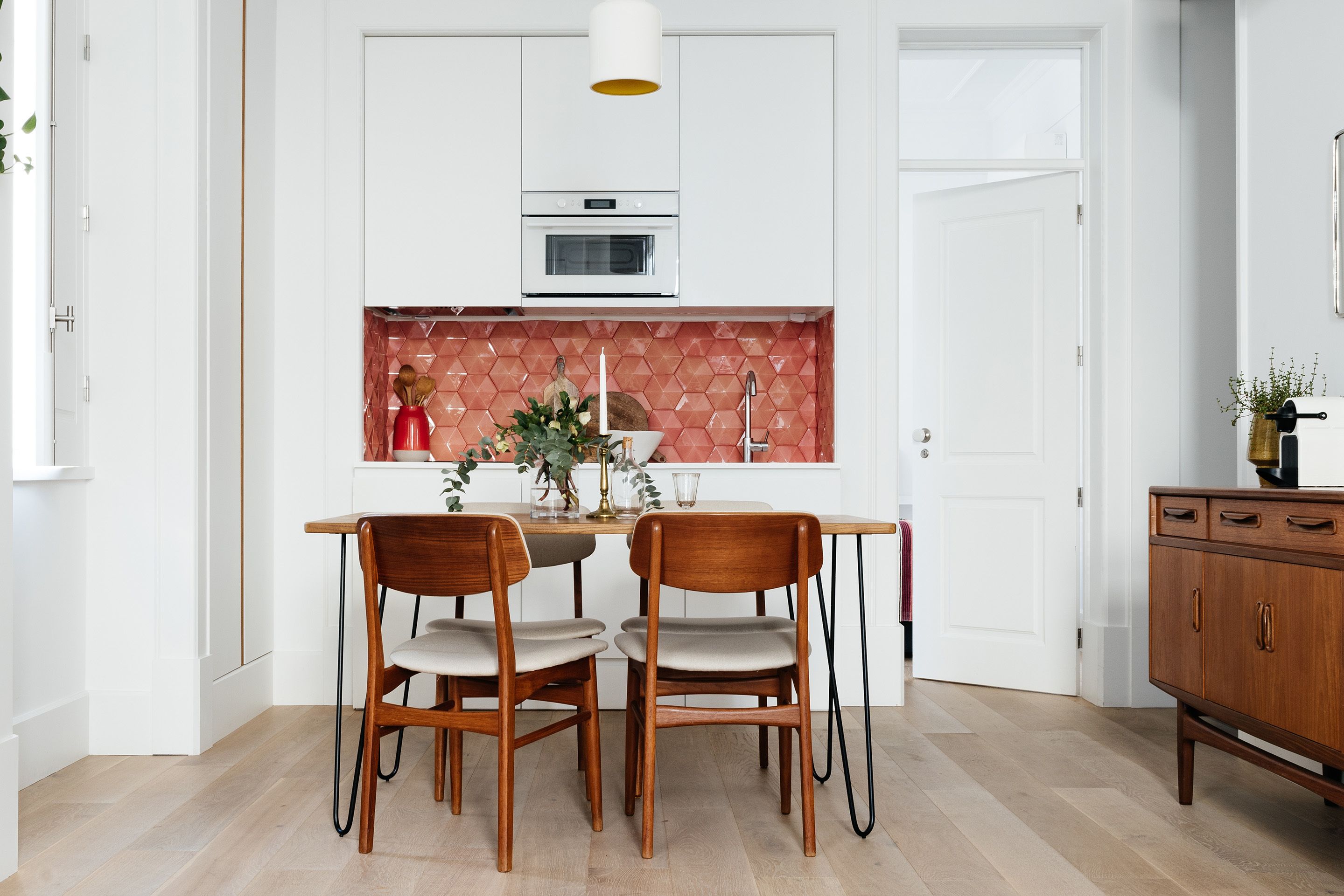 Uma cozinha moderna com armários brancos, uma parede decorada com azulejos vermelhos e uma mesa de jantar de madeira com duas cadeiras.