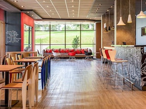 Una cafetería moderna con mesas y sillas de madera, un sofá rojo con cojines, y amplias ventanas que ofrecen vistas al exterior.