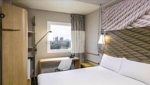 Habitación de hotel con cama, escritorio, silla, lámpara y ventana con vista a edificios.
