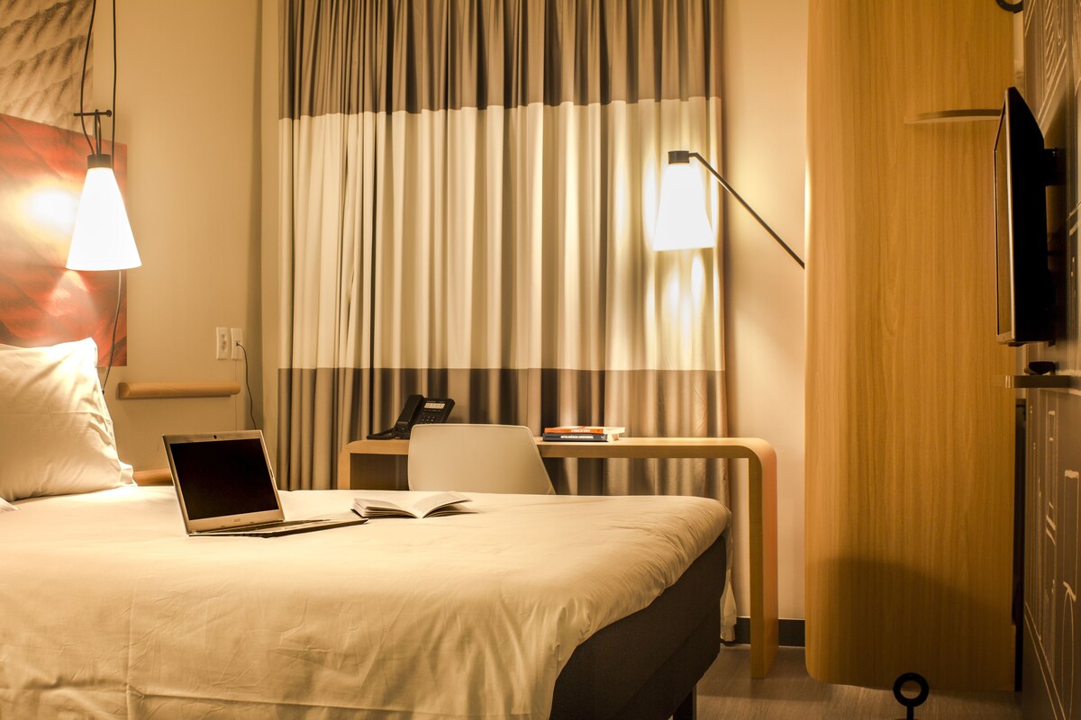 Una habitación de hotel con una cama, un escritorio, una computadora portátil, un teléfono y un televisor.
