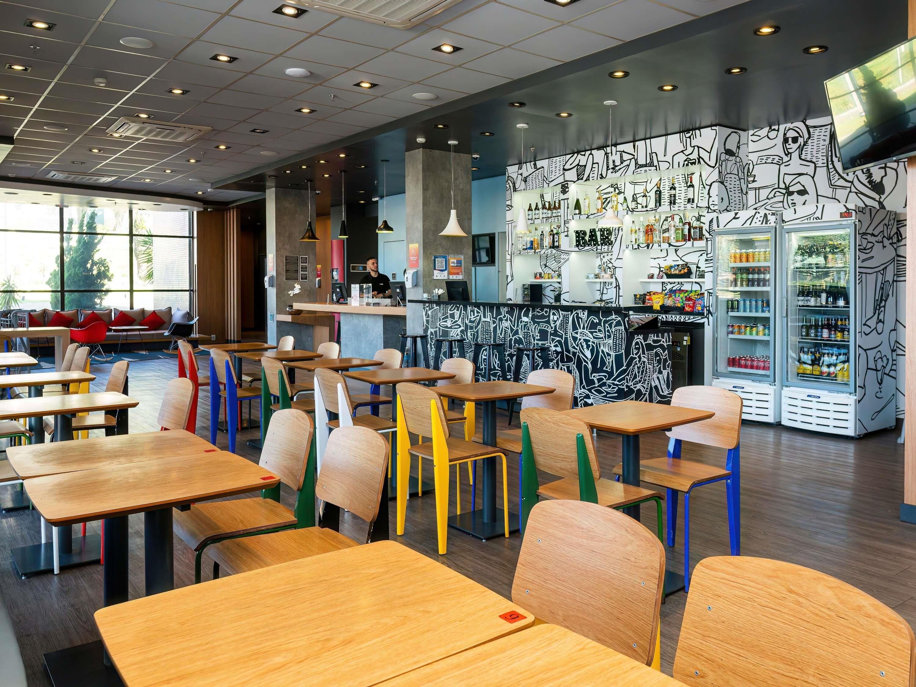 Interior moderno de una cafetería con mesas de madera y sillas coloridas, una zona de bar con pared de grafiti y un amplio ventanal que proporciona luz natural.
