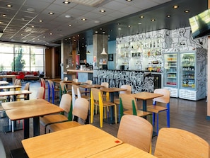Interior moderno de una cafetería con mesas de madera y sillas coloridas, una zona de bar con pared de grafiti y un amplio ventanal que proporciona luz natural.