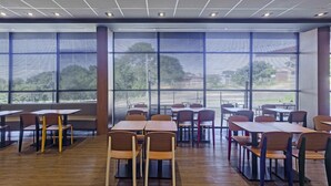 Een moderne eetruimte met houten tafels en stoelen, grote ramen met jaloezieën en een plafond met inbouwverlichting.