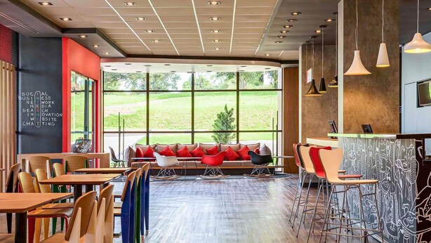 A modern cafe with wooden tables and chairs, a red sofa with cushions, and large windows offering a view of the outdoors. - ibis Guaiba (Guaiba)