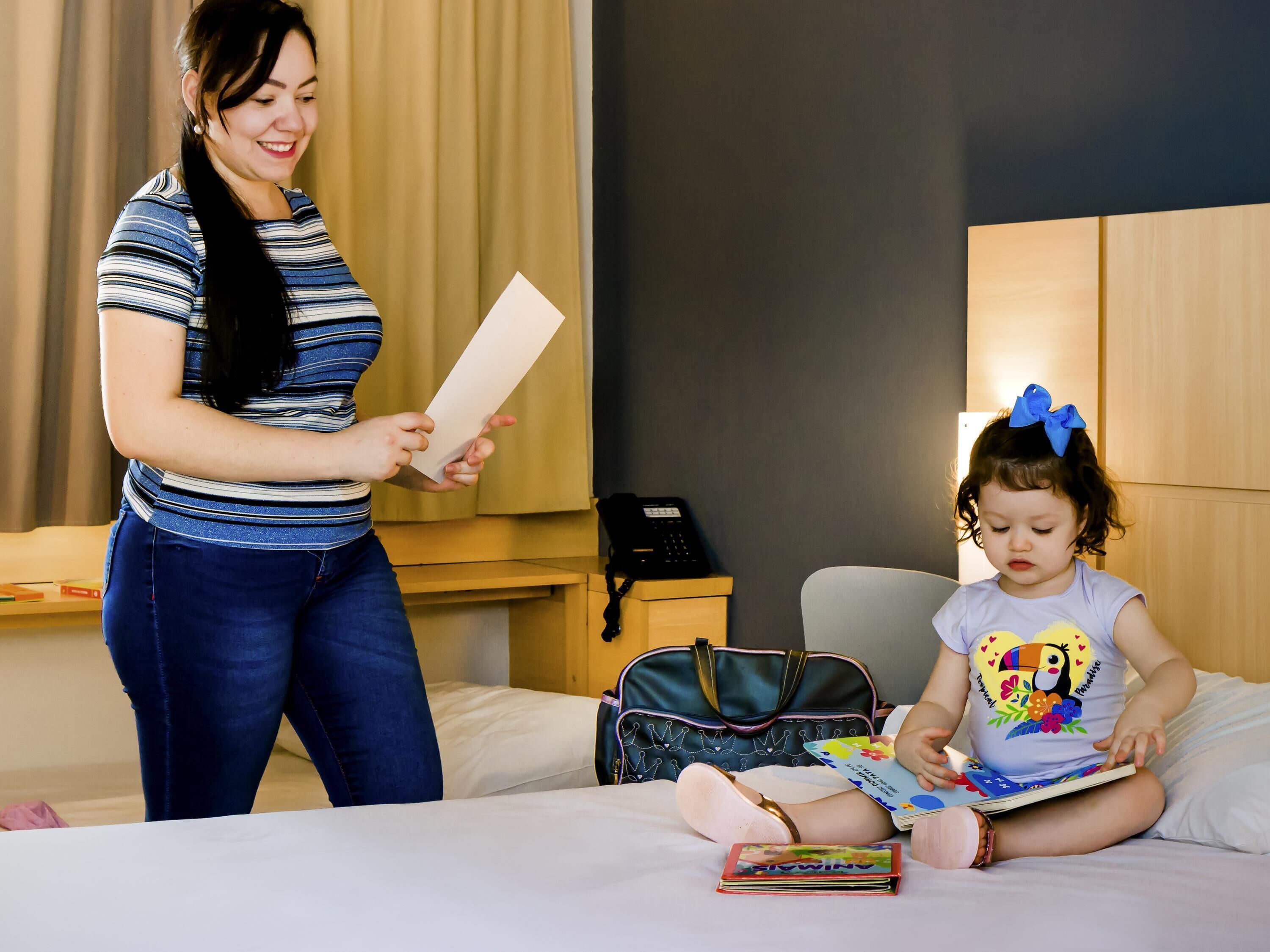 Una mujer y un niño en una habitación de hotel con una cama, una mesita de noche, una lámpara y una maleta.