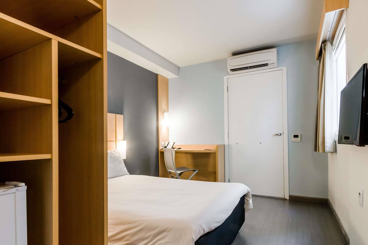 A hotel room with a bed, a desk, a chair, a wardrobe, and an air conditioning unit.