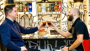 Dos hombres brindando con bebidas en un área de bar con estantes llenos de botellas de licor y un diseño abstracto blanco y negro en la pared.