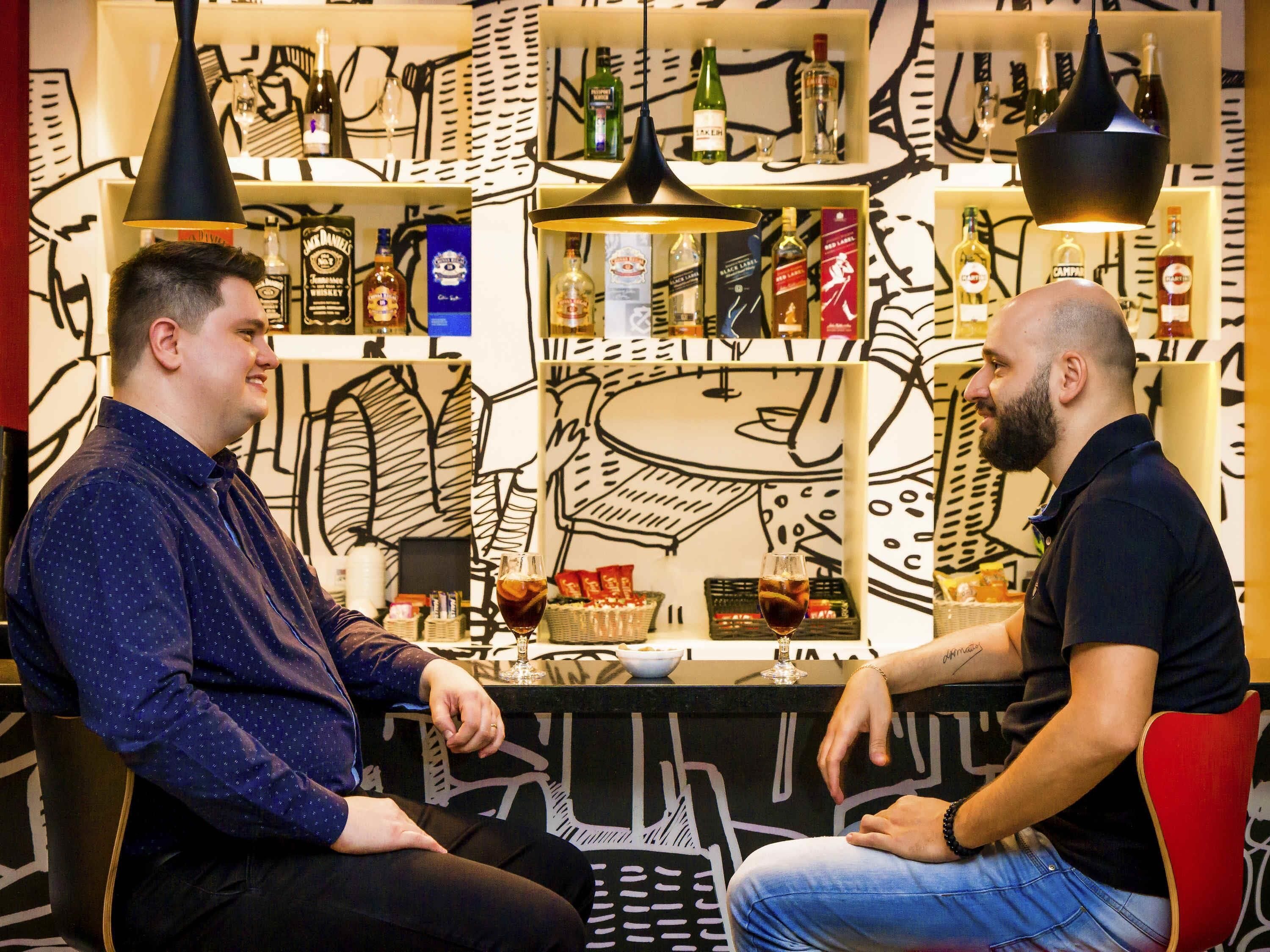 Two men seated at a bar with shelves of liquor bottles behind them.