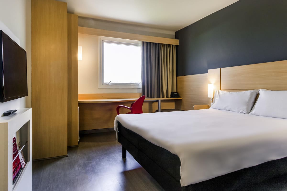 A hotel room with a bed, a desk, a chair, a TV, and a window with curtains.