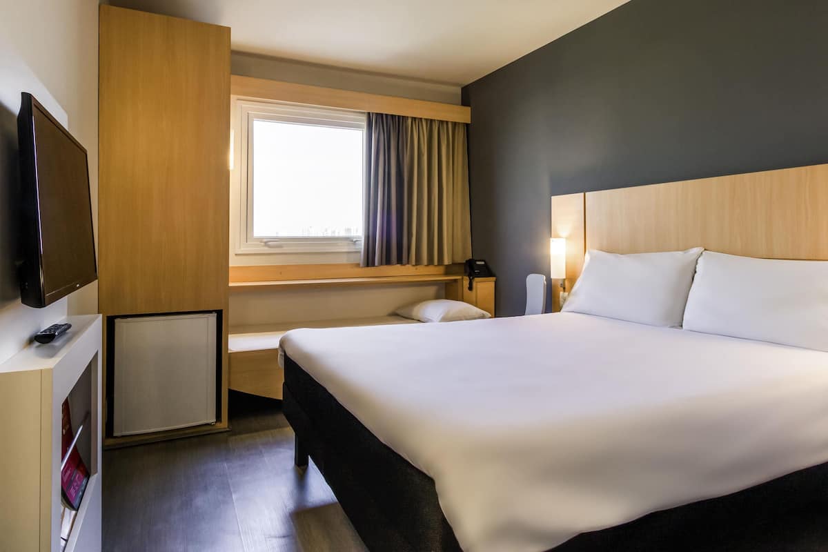 A hotel room with a large bed, a television on a stand, a desk with a phone, and a window with curtains.