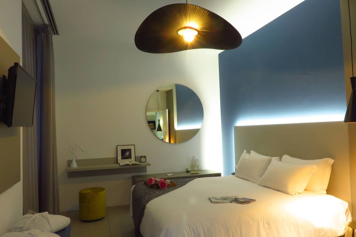 A hotel room with a bed, bedside table, mirror, and a hanging light fixture.