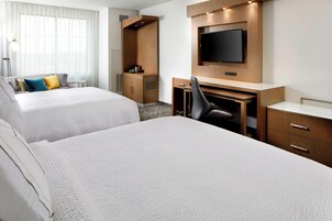 Una habitación de hotel con dos camas, un escritorio con silla, televisor y una mesita de noche con lámpara.