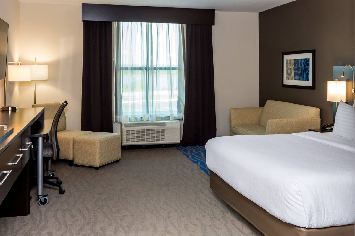 Habitación de hotel con cama, escritorio, silla, unidad de aire acondicionado y ventana con cortinas.