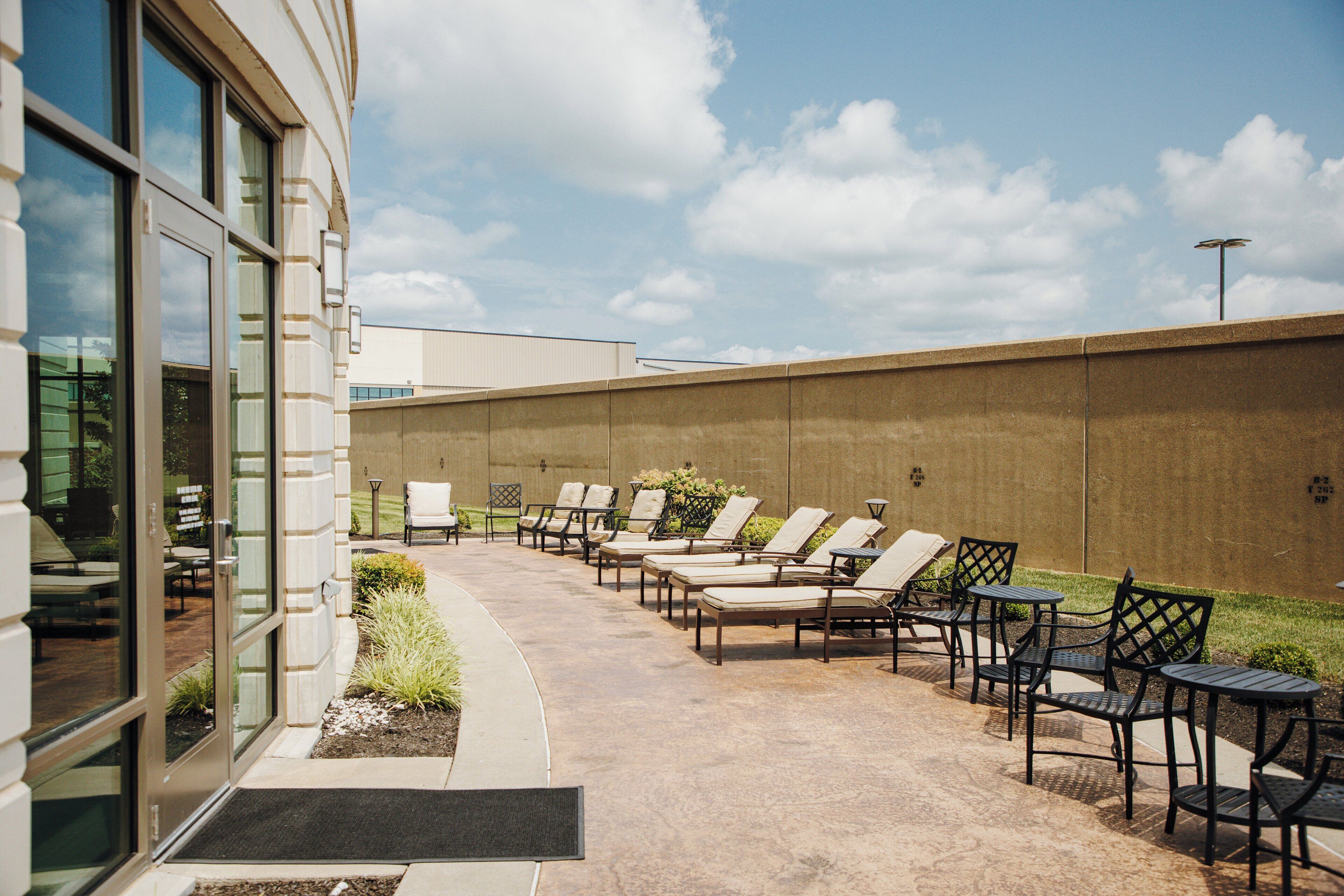 Un espace patio aménagé avec des chaises longues et des tables, un mur en béton et un bâtiment doté de grandes fenêtres.