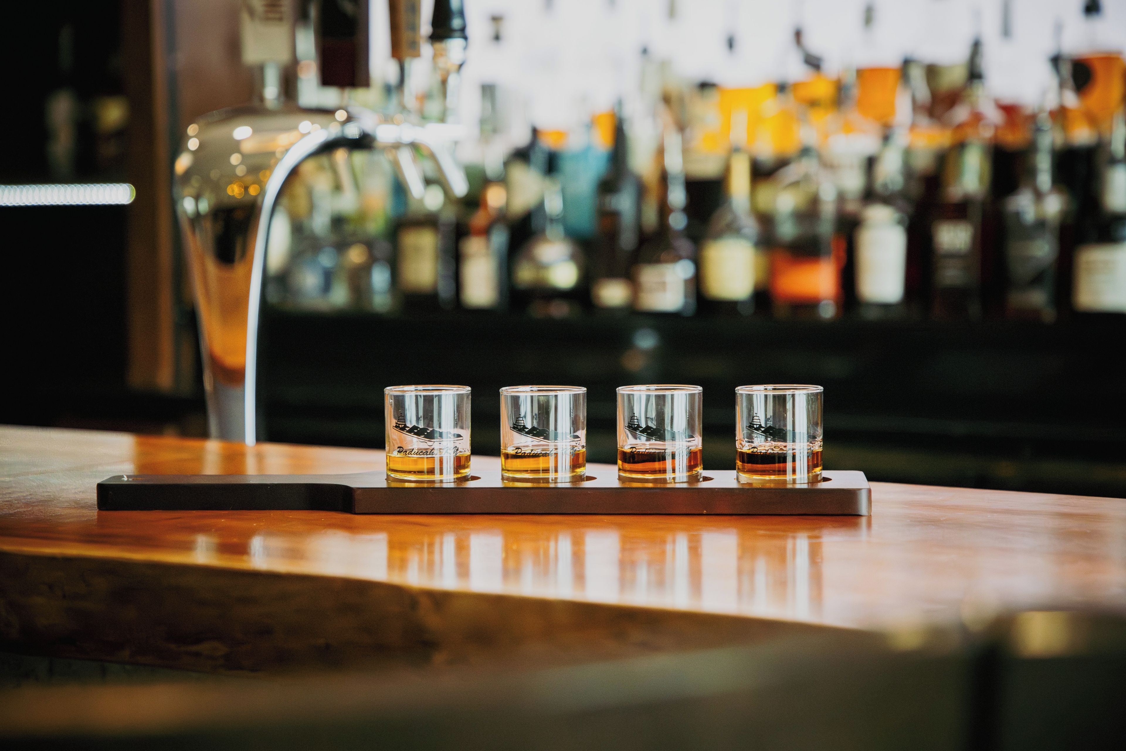Una barra con cuatro vasos de chupito llenos de un líquido dorado, un grifo de metal y un fondo borroso de varias botellas.